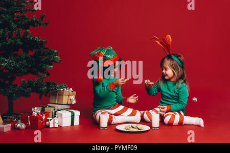 Petites filles, assis près d'un arbre de Noël avec des biscuits et du lait. Deux enfants heureux vêtus de vêtements thème Noël assis sur le plancher à côté d'un ch Banque D'Images