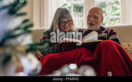 Happy senior man and woman reading a book et souriant. Vieux couple de passer du temps ensemble en lisant un livre assis sur la table à la maison. Banque D'Images