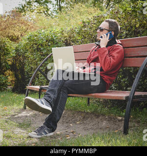 L'homme de travail à l'extérieur toujours connecté avec son smartphone et son fidèle ordinateur portable. jeune mec, le pigiste travailler à l'extérieur dans le parc de l'automne. Un spécialiste informatique Banque D'Images