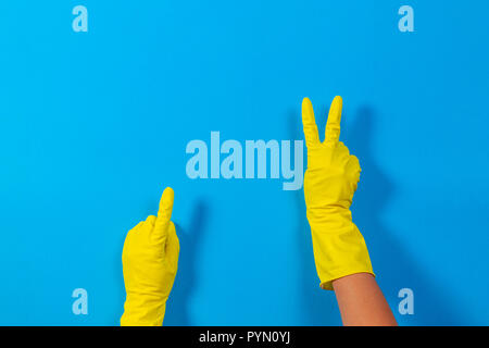 Femme les mains avec des gants en caoutchouc jaune faisant un geste signifiant victoire et pointe vers le haut avec le doigt, fond bleu. Banque D'Images