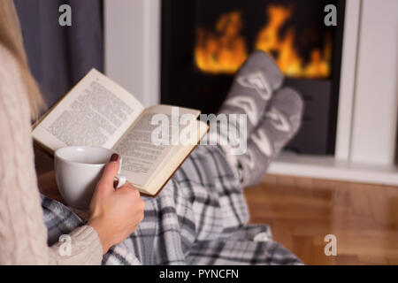 Jeune fille devant la cheminée reading book et réchauffement pieds sur le feu et les jambes sont couverts avec une couverture, dans la main est titulaire d'tasse de thé chaud. Hiver et froid Banque D'Images