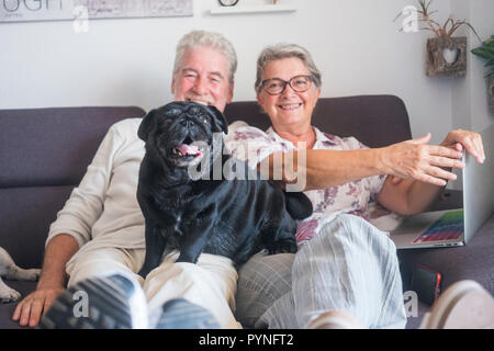 Couple de personnes adultes matures caucasienne heureux homme et femme assise sur le canapé avec un ordinateur portable et black pug dog drôle sur lui à la recherche de l'appareil photo Banque D'Images