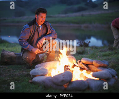 Man crouching à côté d'un feu de camp près d'une rivière. Banque D'Images