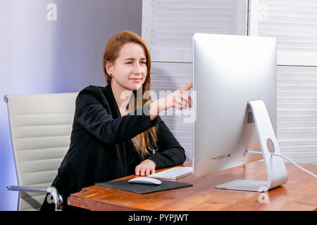 Portrait of happy young businesswoman réussie au bureau. Elle s'assoit à la table et les points à l'écran. Le vendredi noir ou Cyber lundi. La sélection d'i Banque D'Images