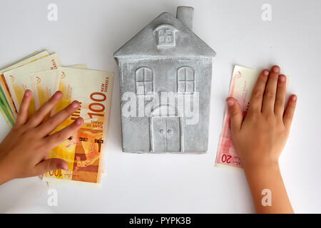 La composition créative de pierre maison maquette et kid holding pile de nouveau shekel israélien sur fond blanc Banque D'Images