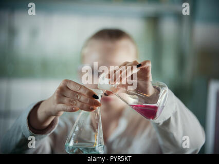 Laboratoire chimique. Jeune femme blonde sur le point de verser un liquide rose dans le bleu. Banque D'Images