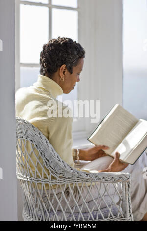 Mid-adult woman sitting in a wicker chair lire un livre dans une chambre ensoleillée. Banque D'Images