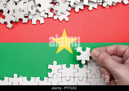 Burkina Faso drapeau est représenté sur une table sur laquelle la main de l'homme plie un puzzle de couleur blanche. Banque D'Images