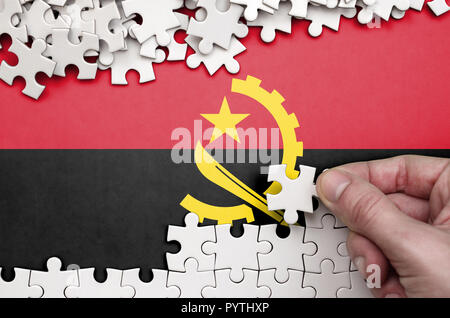 Drapeau de l'Angola est représenté sur une table sur laquelle la main de l'homme plie un puzzle de couleur blanche. Banque D'Images