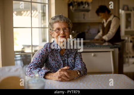 Portrait d'une femme âgée assise à sa table de cuisine tandis que sa fille aide à faire la vaisselle. Banque D'Images