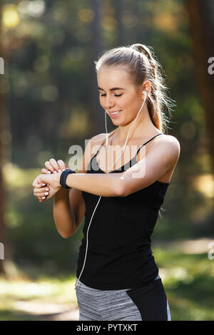 Long Sportish-haired young woman wearing black shirt debout dans le parc ensoleillé, écouter la musique et à la recherche sur la montre de sport, mode de vie sain et les gens concept Banque D'Images