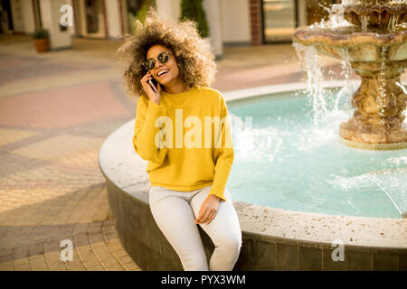 Aftican american jeune femme est assise près de la fontaine et les pourparlers sur le smartphone. Banque D'Images