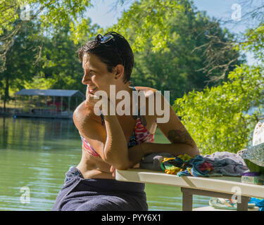 Happy young woman en maillot de bain assise seule à la table smiling à par-dessus son épaule au lac en été Banque D'Images