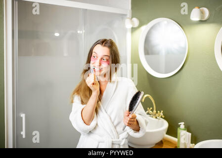 Jeune et belle femme en peignoir se brosser les dents dans la salle de bains Banque D'Images