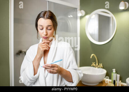 Tristes et inquiets femme en peignoir en attente d'une suite à la recherche sur un test de grossesse stick dans la salle de bains Banque D'Images