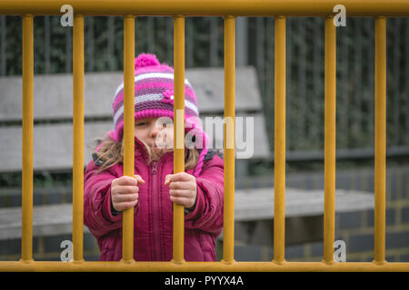 Peu cute young girl in pink hat et veste debout derrière des portes métalliques menant à l'aire de jeux Banque D'Images