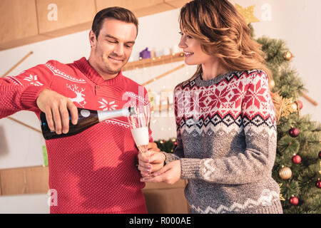 Couple heureux à Noël chandails, smiling mari pouring champagne pour femme Banque D'Images