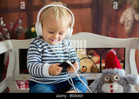 Sweet bébé garçon avec des écouteurs, l'écoute de la musique, assis sur banc rustique, décoration de Noël derrière lui. L'enfance, pour les enfants, de la technologie et de Chris Banque D'Images