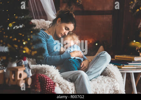 Mère d'allaiter son petit garçon assis dans un fauteuil confortable près de l'arbre de Noël, l'hiver Banque D'Images