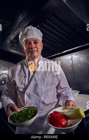 Chef cuisinier professionnel dans un tablier blanc et de cuisiniers hat prepairing délicieux repas sur une cuisine avec bol plein de viande Banque D'Images