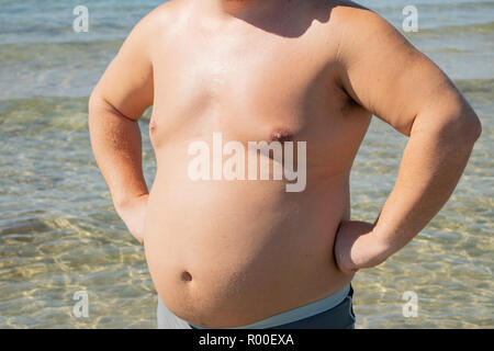 Un gros homme en short se dresse sur la rive de la mer. Soleil et locations Banque D'Images