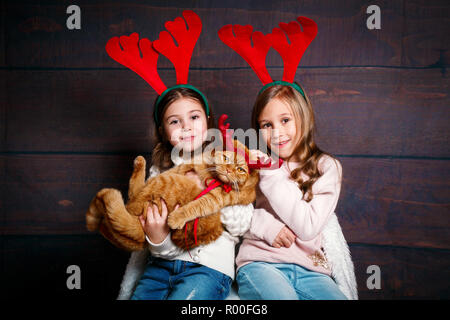 Deux drôles de petites filles souriant avec chat rouge à cornes de cerf sur fond de bois. Joyeux Noël et Bonne Année . Banque D'Images