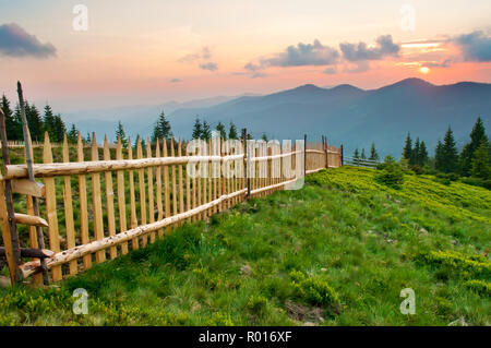 Les épinettes sur pente verdoyante contre sommets de montagne couverte de plusieurs nuages à coucher du soleil. Diagonale de clôture en bois. L'été chaud evenin Banque D'Images