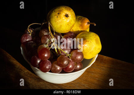 Bol blanc sur marron contenant des raisins de table et trois poires isolé sur fond noir. Banque D'Images