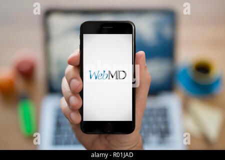 Un homme se penche sur son iPhone qui affiche le logo WebMD, en restant assis devant son ordinateur 24 (usage éditorial uniquement). Banque D'Images