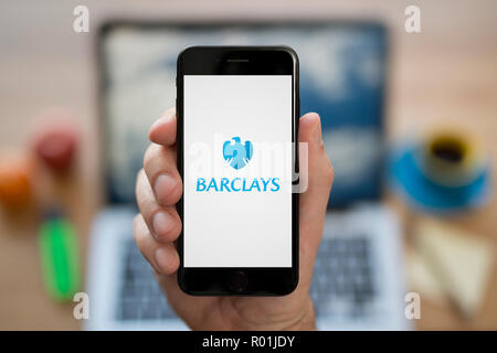 Un homme se penche sur son iPhone qui affiche le logo de la Barclays, tandis qu'assis à son bureau de l'ordinateur (usage éditorial uniquement). Banque D'Images