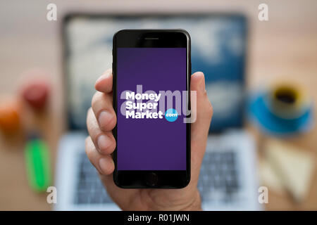 Un homme se penche sur son iPhone qui affiche le logo du supermarché de l'argent, alors qu'assis à son bureau de l'ordinateur (usage éditorial uniquement). Banque D'Images