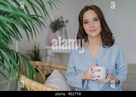 Photo de jolie femme avec look attrayant, vêtu de vêtements nationaux occasionnels, détient avec mug blanc café ou thé aromatique, est assis sur des meubles en rotin Banque D'Images