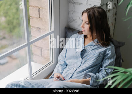 Coup horizontal d'une jeune femme est assise sur le rebord de la fenêtre, vêtu de vêtements décontractés, utilise des smart phone et écouteurs, écoute la musique calme tandis que lo Banque D'Images