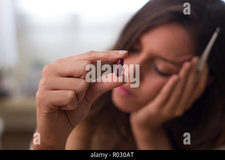 Part de mauvais jeune brunette woman holding un comprimé de médicament contre la fièvre, maux de tête, les maladies chroniques et thermomètre blanc ; avec un visage triste, fermé avec Banque D'Images