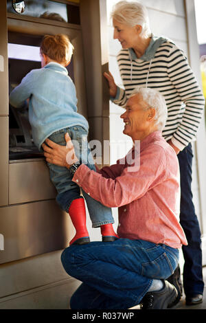 Petit garçon essayant de se retirer de l'argent de la cash machine avec l'aide de ses grands-parents. Banque D'Images