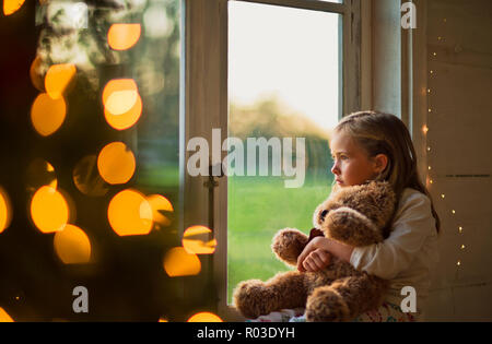 Jeune fille en regardant par la fenêtre, tenant son ours en peluche. Banque D'Images