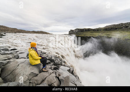 Femme en imperméable jaune assis debout sur le bord de la falaise à côté de la cascade Dettifoss dans Vatnayokull, parc national de l'Islande. Banque D'Images