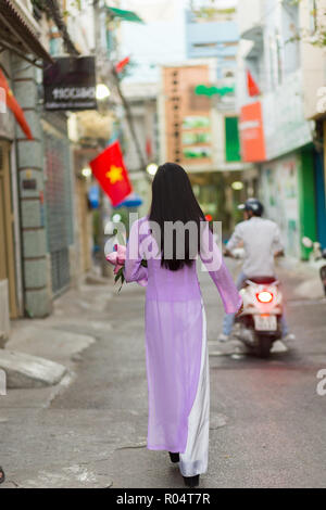 Vietnamese woman walking in Ho Chi Minh street port Ao Dai costume traditionnel, portant des fleurs de lotus avec le drapeau du Vietnam en arrière-plan . Banque D'Images