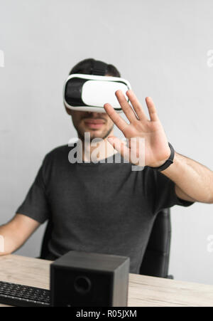 Jeune homme portant des lunettes et un casque de réalité virtuelle assis au bureau contre l'ordinateur. La liaison, de la technologie, nouvelle génération. L'homme essaie de toucher des objets ou de contrôle avec un VR main. Banque D'Images