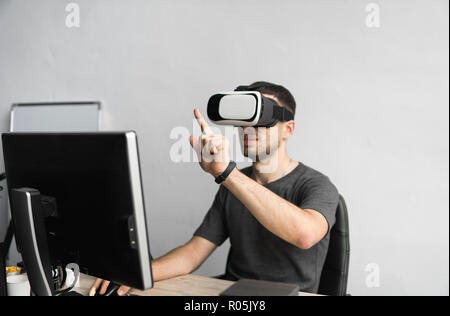 Jeune homme portant des lunettes et un casque de réalité virtuelle assis au bureau contre l'ordinateur. La liaison, de la technologie, nouvelle génération. L'homme essaie de toucher des objets ou de contrôle avec un VR main. Banque D'Images