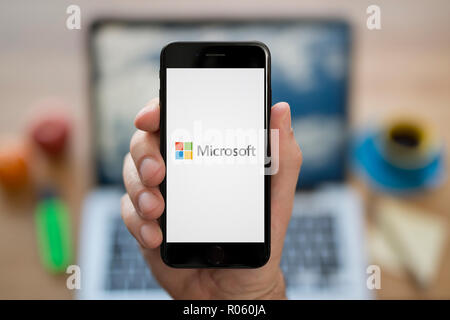 Un homme se penche sur son iPhone qui affiche le logo de Microsoft, tout en s'assit à son bureau informatique (usage éditorial uniquement). Banque D'Images