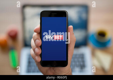 Un homme se penche sur son iPhone qui affiche le logo Sky Bet, tandis qu'assis à son bureau de l'ordinateur (usage éditorial uniquement). Banque D'Images