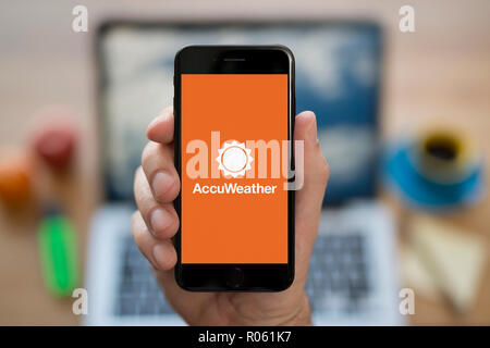 Un homme se penche sur son iPhone qui affiche le logo Accuweather, tandis qu'assis à son bureau de l'ordinateur (usage éditorial uniquement). Banque D'Images