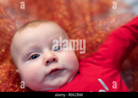 Chubby blue eyed bébé dans un lit bébé Noël allongé sur un fond orange scintillant dans un close up face portrait Banque D'Images