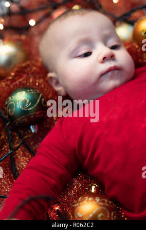 Cute little baby avec boules de Noël coloré couché dans un lit bébé sur un tissu rouge scintillant dans une saison de vacances concept Banque D'Images