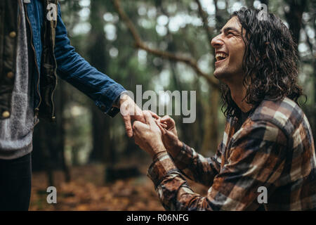 Smiling young man putting bague de fiançailles sur la main de femme à l'extérieur sous la pluie. Cheerful guy faire proposition de mariage de petite amie à parc. Banque D'Images