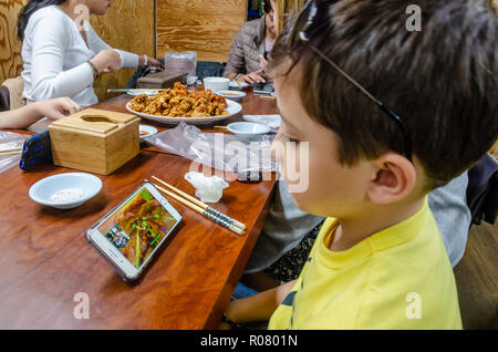 Un enfant regarde une vidéo Youtube sur un téléphone mobile en restant assis à la table à manger dans un restaurant pour le divertissement. Banque D'Images