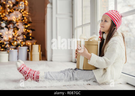 Portrait de femme reposant sur le côté enfant porte chandail blanc, un pantalon et des chaussettes chaudes, englobe des cadeaux emballés, est assis sur le plancher en chambre douillette, admire Nouveau Banque D'Images