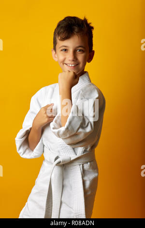 Heureux petit garçon en kimono blanc de fighter dans la position debout smiling at camera sur fond jaune Banque D'Images