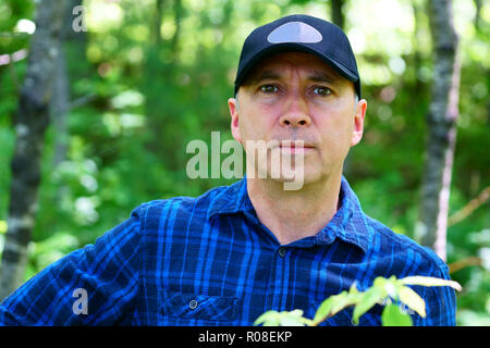 Un sérieux man est à la recherche dans l'appareil photo pour un portrait lors d'une randonnée dans la forêt portant une chemise à carreaux bleu et un chapeau noir. Banque D'Images